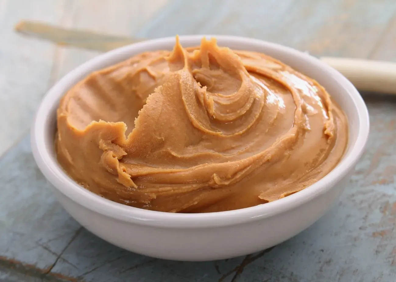 A small white bowl filled with smooth, thick peanut butter, its glossy surface swirled into soft peaks on a rustic blue wooden table.