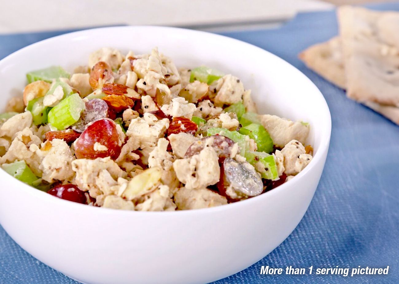 Chicken-Flavored Plant-Based Protein Pouch A close-up of a prepared chicken salad sandwich on a toasted croissant, topped with fresh lettuce, served on a white plate alongside a glass of iced tea and a bowl of fresh grapes.