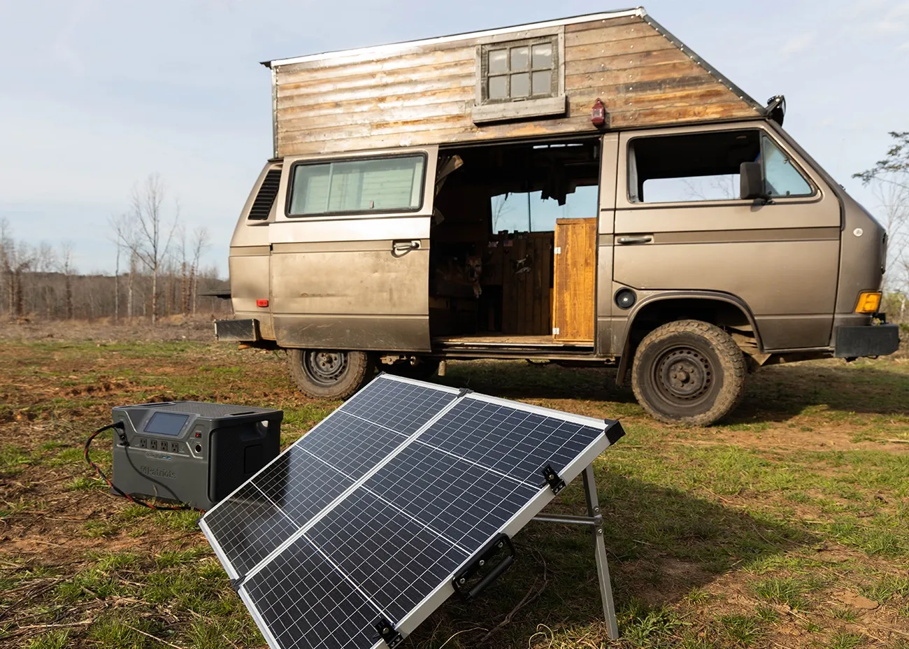 A retro camper van with a wooden pop-top roof is parked on grassy land with its side door open. In front of it, a portable solar panel and 4Patriots power station are set up, showcasing off-grid energy for van life and outdoor living.