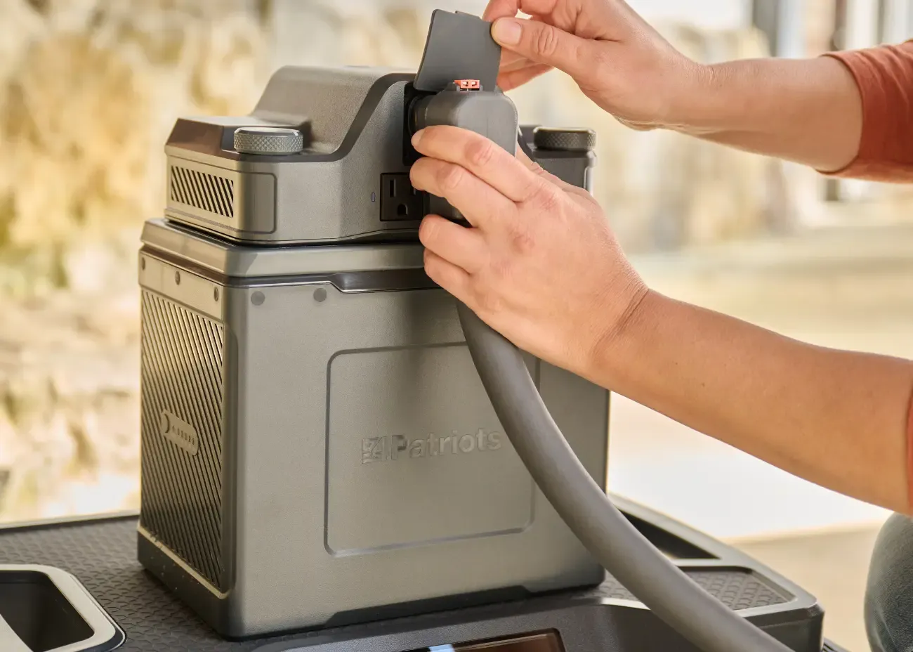 A person’s hands are shown connecting a charging cable to a portable power station branded “4Patriots.” The sturdy, dark gray unit features an outlet port and textured sides, suggesting it’s designed for off-grid or emergency use.