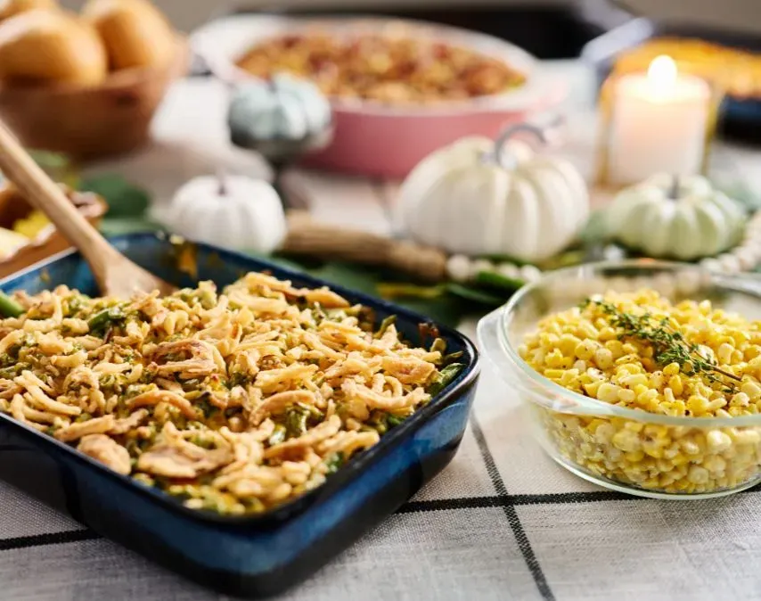 A festive table setting with a blue baking dish of green bean casserole topped with crispy onions beside a glass bowl of seasoned corn, surrounded by fall decorations and a lit candle.