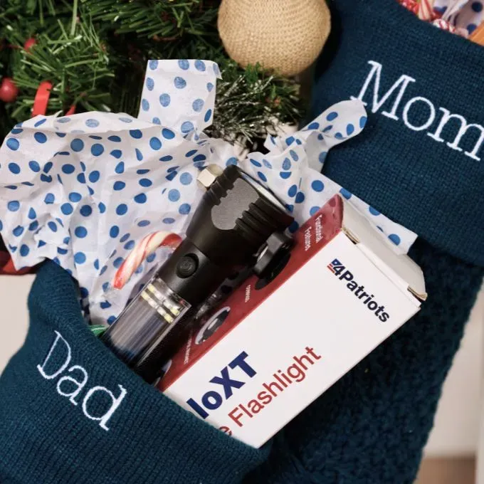 Christmas stockings labeled “Dad” and “Mom” filled with gifts including a 4Patriots HaloXT flashlight in its box, candy canes, and polka-dotted tissue paper.