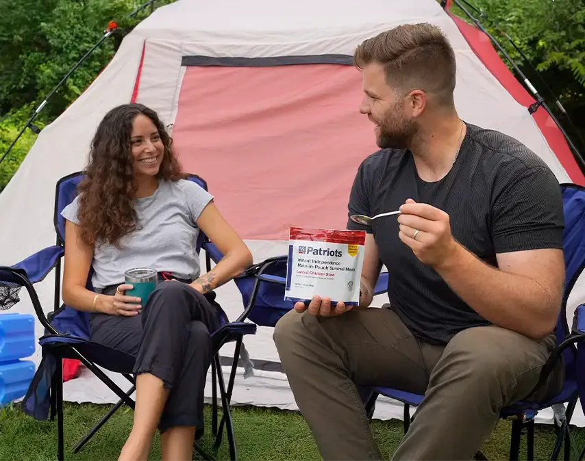 Two people sit outside a pink and white camping tent, smiling and enjoying a moment together. The man is holding a pouch labeled “4Patriots Instant Independence Meal-In-Pouch Survival Meal – Creamy Chicken Rice."