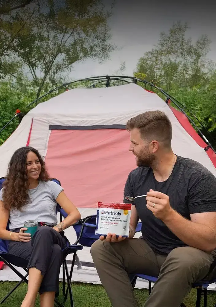 Two people sit outside a pink and white camping tent, smiling and enjoying a moment together. The man is holding a pouch labeled “4Patriots Instant Independence Meal-In-Pouch Survival Meal – Creamy Chicken Rice."