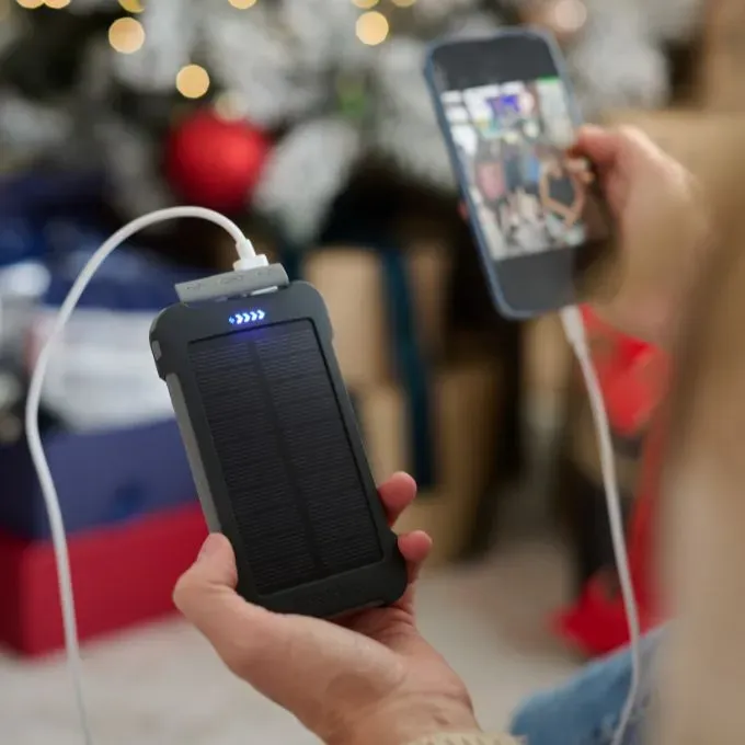 A person holding a solar power bank charging a smartphone via USB cable, with a lit Christmas tree and gifts blurred in the background.
