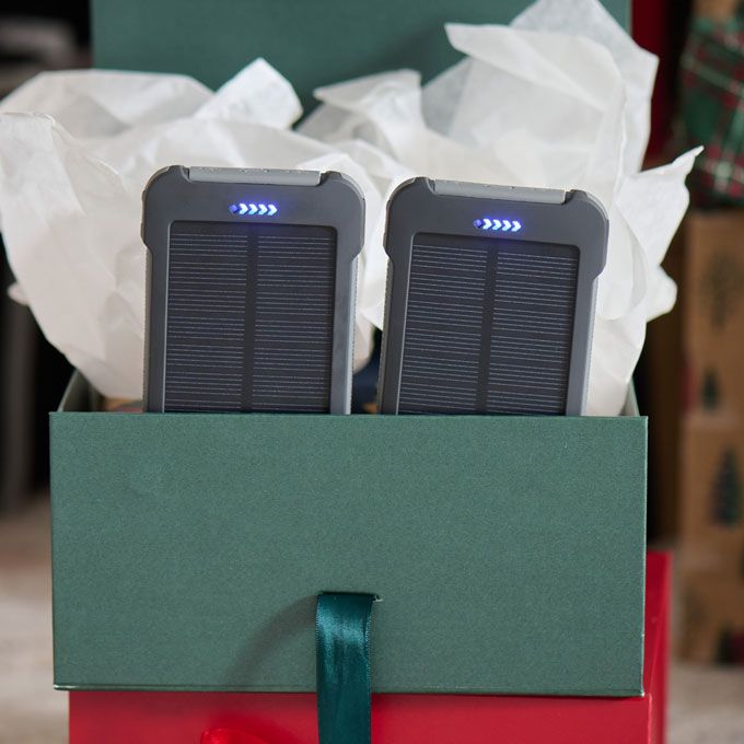 A dark‑green gift box with white tissue paper displays two black Patriot Power Cell-CX Power Banks, each featuring a visible blue LED indicator light. The holiday-themed packaging suggests it is ready for gift-giving.
