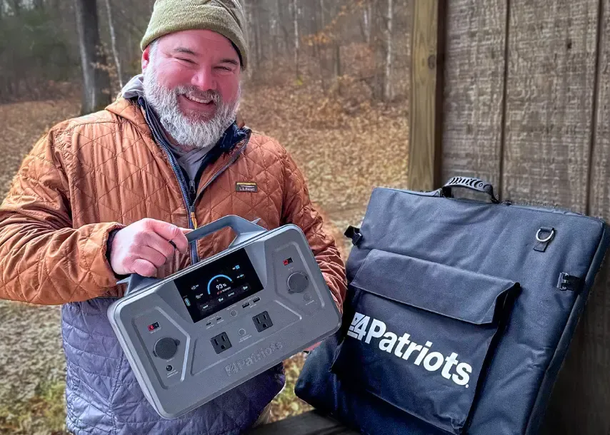 A smiling man in an orange jacket holds a 4Patriots portable power station next to its carrying case, standing outdoors near a wooden wall with a forest in the background.