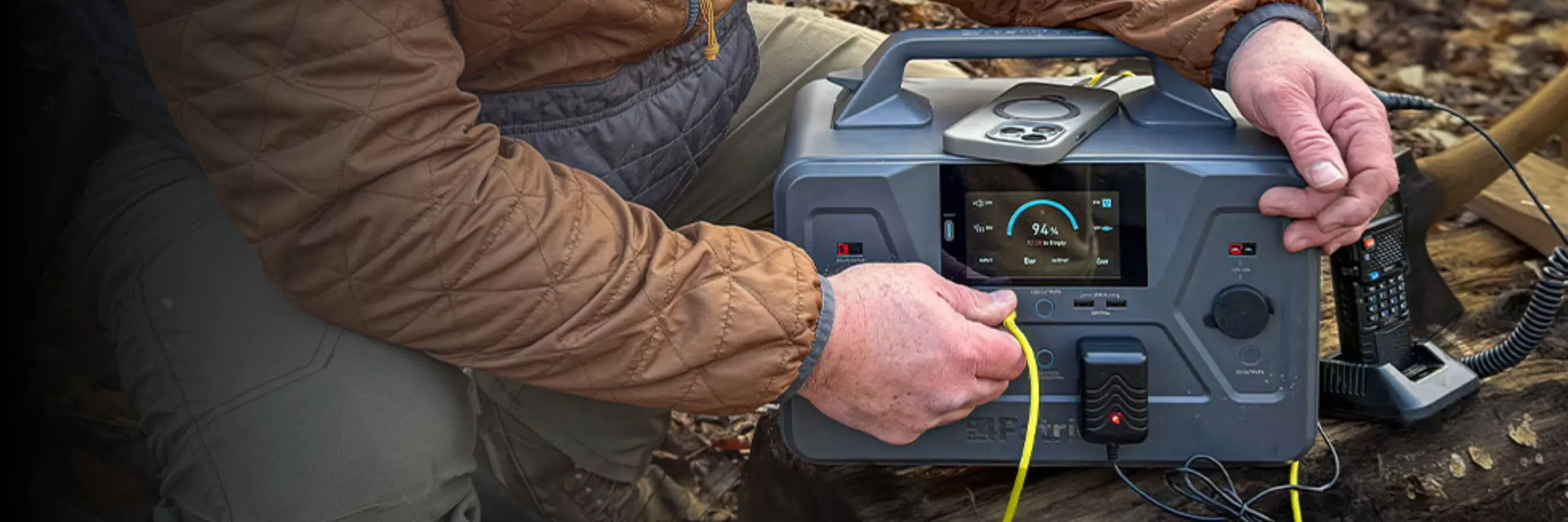 A person in outdoor gear uses a 4Patriots portable power station 1200 on the ground, with the display showing a 94 percent charge. The unit is powering a device with a yellow cable and sits next to a handheld radio, suggesting off-grid or emergency use.