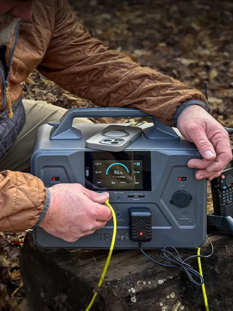 A person in outdoor gear uses a 4Patriots portable power station on the ground, with the display showing a 94 percent charge. The unit is powering a device with a yellow cable and sits next to a handheld radio, suggesting off-grid or emergency use.