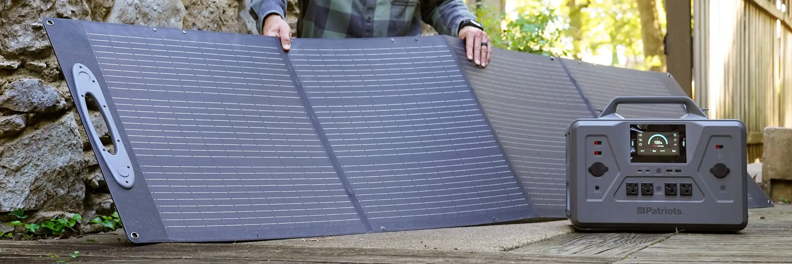 Patriot Power Solar Generator 2500X & 2 100W Solar Panels A woman sets up portable solar panels on a grassy clearing while a man sits nearby on a wooden bench holding a tablet. In the foreground, a 4Patriots solar generator is connected to the panels, with autumn trees in the background.