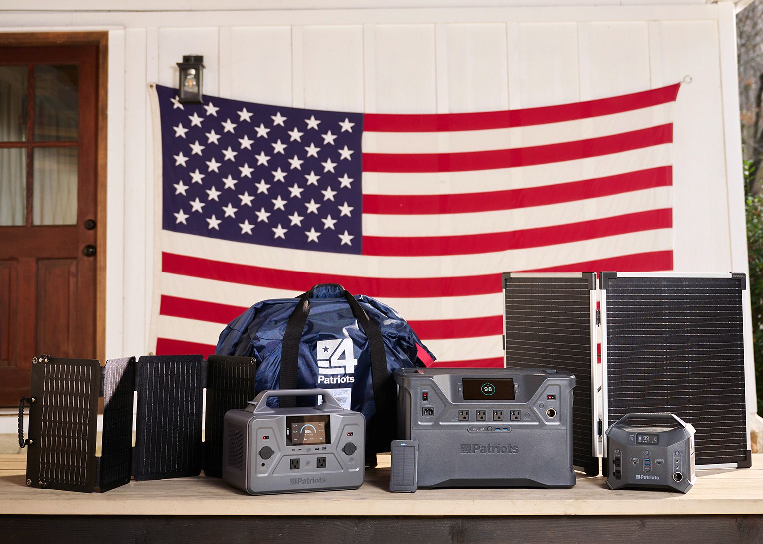 4Patriots Family of Solar Generators Display of multiple 4Patriots portable solar generators and foldable solar panels arranged on a wooden porch in front of a large American flag.