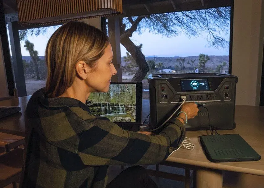 A woman sits at a table plugging a device into a fully charged 4Patriots portable power station, with a laptop and tablet nearby and a scenic desert view outside the window.