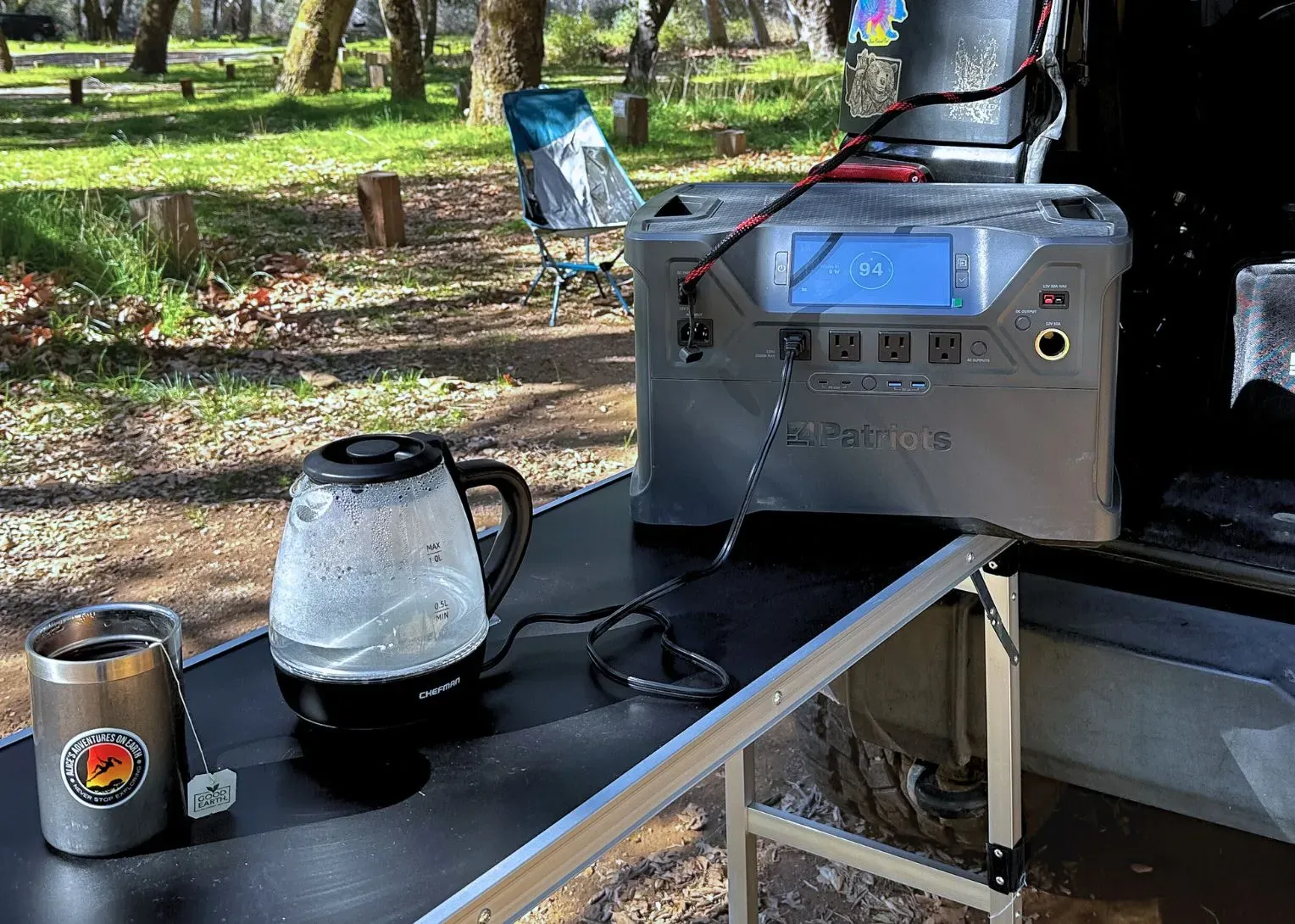 A 4Patriots portable power generator set up at a campsite, powering a coffee pot while connected to a vehicle. The display shows 94% charge, with a camping chair and trees in the background.