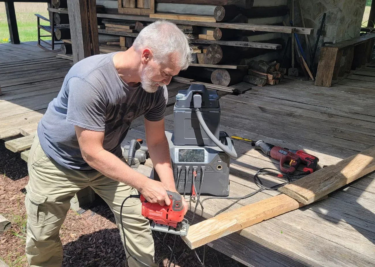 A man uses a power saw connected to a 4Patriots generator with an external battery pack. The setup is on a wooden porch, showing the generator powering heavy-duty tools for off-grid work.