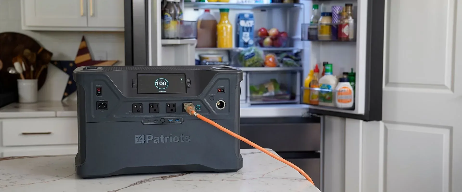 A 4Patriots portable power station sits on a kitchen counter with an orange power cord plugged into it, supplying backup power to an open refrigerator stocked with groceries. The digital display on the power station shows a full 100% charge, emphasizing its emergency preparedness role.