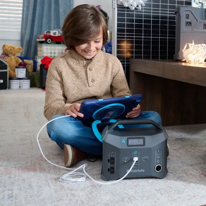 A child sitting on the floor playing with a tablet connected to a 4Patriots Sidekick portable power station, with presents and a solar panel behind them.
