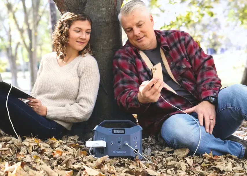A man and woman sit against a tree in autumn leaves while charging devices from a 4Patriots portable power station placed on the ground between them.
