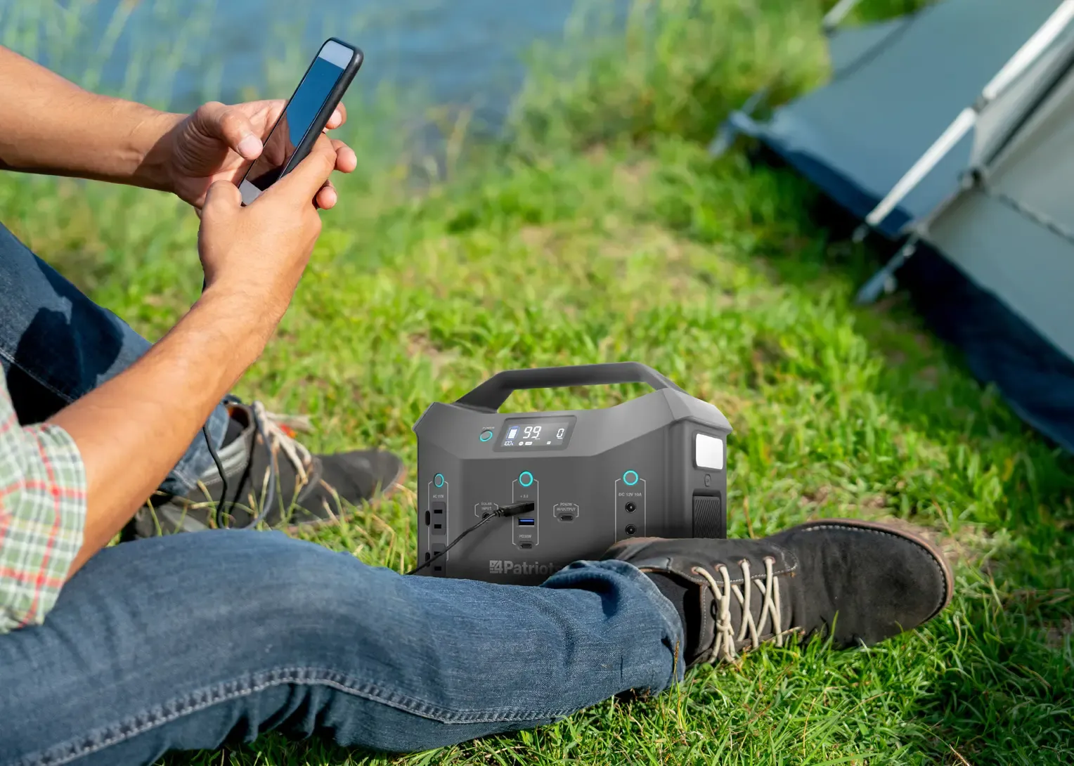 Close-up of a person charging a phone with a 4Patriots Sidekick portable power station sitting on grass.