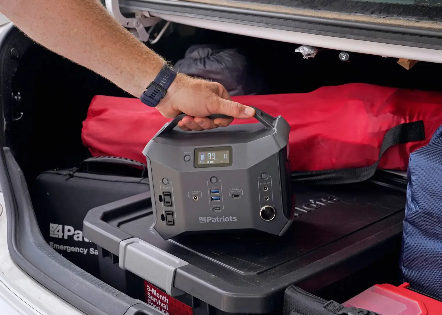 Close-up of a person loading a 4Patriots Sidekick portable power station into a vehicle trunk, surrounded by emergency gear including a labeled 3-Month Survival Food Kit. The power station displays a 99% charge, emphasizing its readiness for off-grid or emergency use.