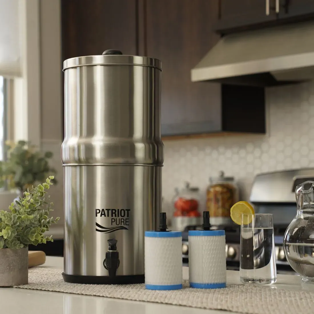 Stainless steel Patriot Pure water filter system displayed on a kitchen counter, with two replacement filters, a glass of water with a lemon slice, and a pitcher, showcasing the ultimate water filtration system for clean, fresh drinking water.