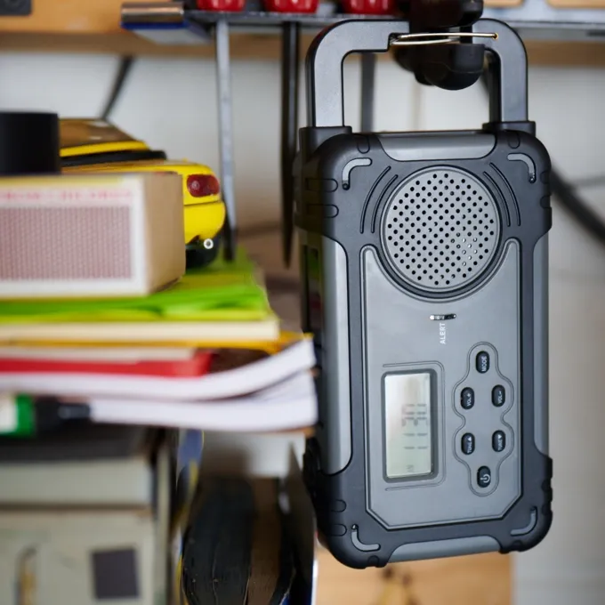 A gray and black Liberty Band Emergency Radio hangs by its built-in handle from a shelf in a workshop. The rugged, compact radio features a front-facing speaker, a digital display screen, and tactile buttons for volume, tuning, and power. The surrounding environment includes workshop tools and stacked papers, highlighting its use as a durable, portable device for emergency preparedness or everyday utility.