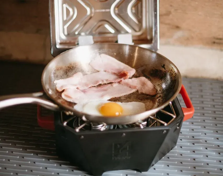 A frying pan with bacon and a sunny-side-up egg cooks on a compact 4Patriots portable stove, with the stove lid propped open and red handles visible.