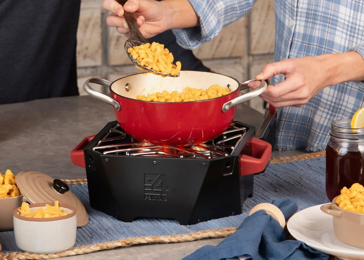 A red pot of macaroni and cheese is being served from a 4Patriots RescueHeat Stove & Header with two people dishing out the meal.