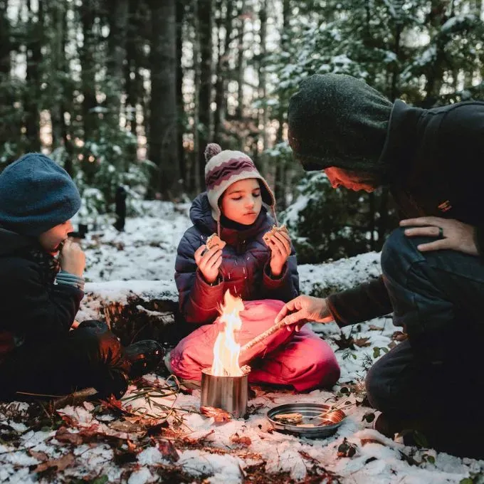 A man and two children gathered around a small fire in a snowy forest, roasting marshmallows and eating s’mores while bundled in winter clothes.