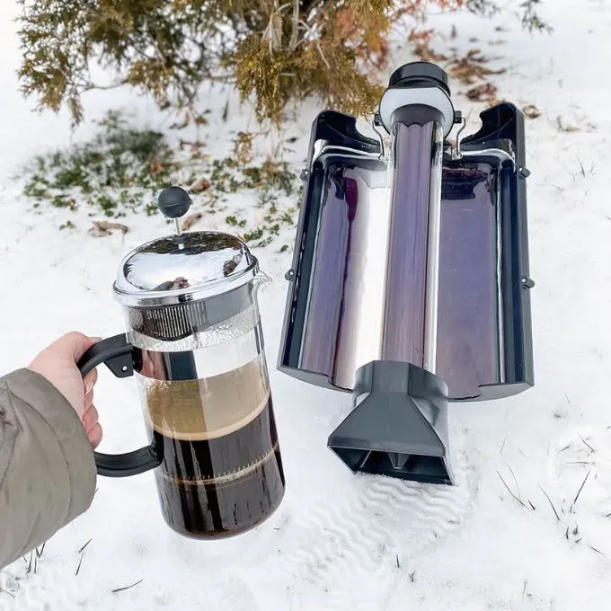 A hand holding a French press full of coffee beside a reflective tube-style Sun Kettle solar cooker on snow-covered ground, with trees in the background.