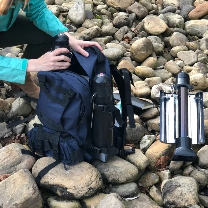 Sun Kettle Solar Water Heater Person packing a 4Patriots Sun Kettle into a navy blue tactical backpack on a rocky riverbank, with another Sun Kettle already set up nearby. The scene highlights the kettle’s portability and readiness for outdoor use.