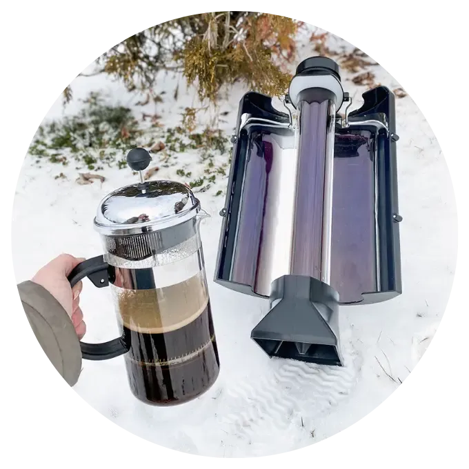 Sun Kettle Solar Thermos A person holds a glass French press filled with coffee next to a Sun Kettle Solar Thermal Cooker resting in the snow. The solar kettle features a long, dark evacuated glass tube centered between two reflective metallic wings that capture sunlight. The background shows a snowy outdoor setting with sparse winter vegetation.