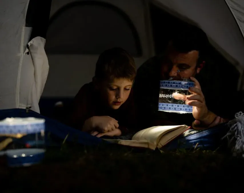 A father and son read a book together inside a tent at night, softly illuminated by a glowing 4Patriots lantern.