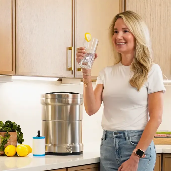 Smiling woman in a modern kitchen holding a glass of lemon water beside the 4Patriots Ultimate Water Filtration System with fresh lemons and a small plant on the counter.